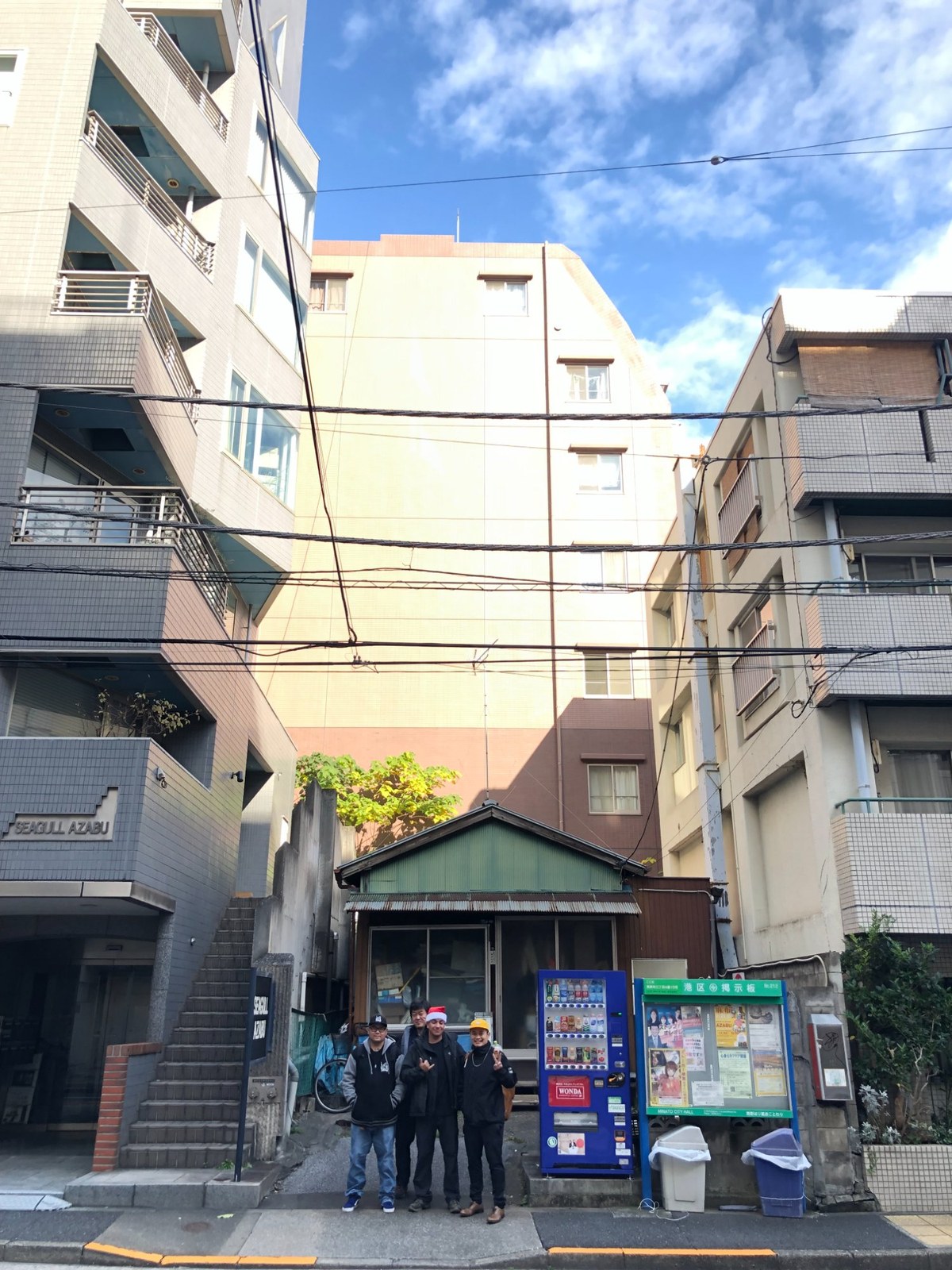 Dino and three friends standing together on a street in Azabu, Tokyo, in front of an old timber shopfront beside a vending machine and a ward noticeboard.